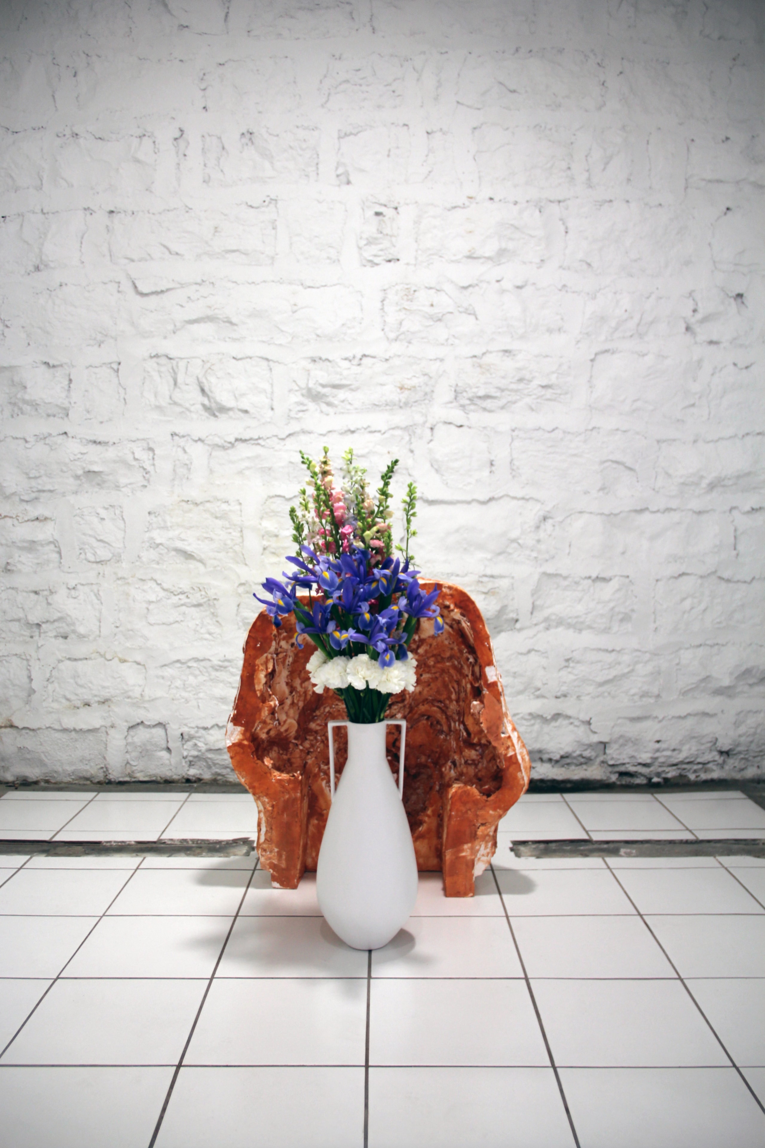 Flower arrangement in a white vase before a terracotta sculptural form and white brick wall
