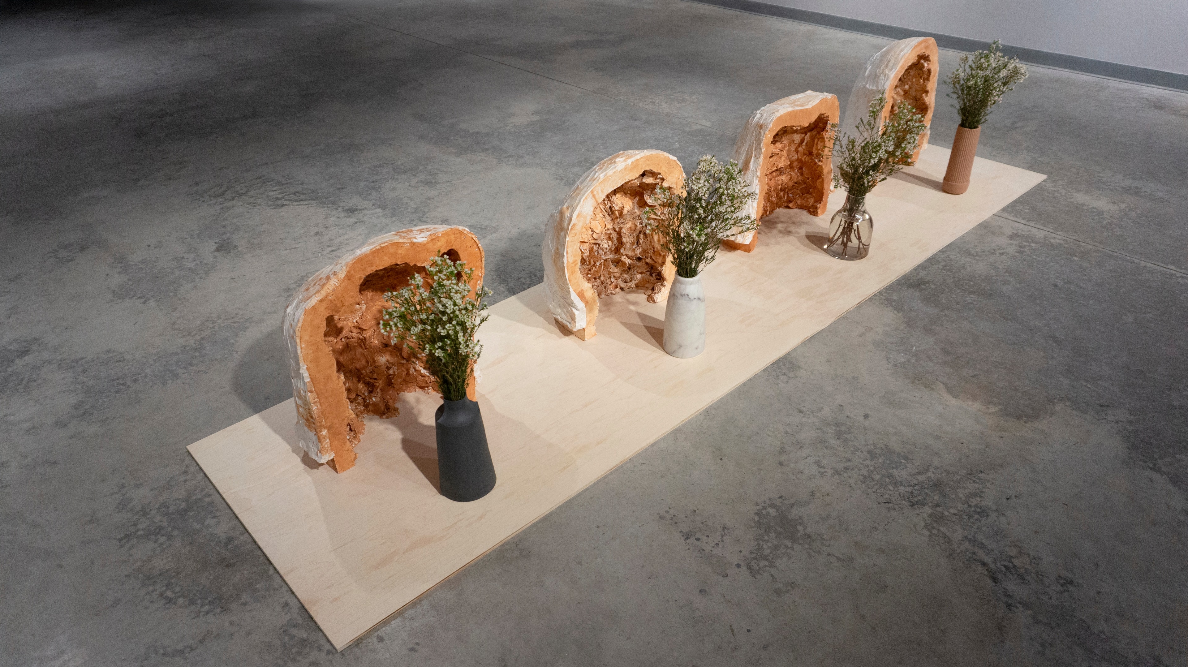 Installation view of sculptural stone forms, vases, and flowers arranged on a pale wooden platform